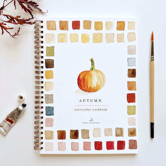 A watercolor workbook with a pumpkin illustration on the cover, surrounded by watercolor paint tubes and a paintbrush. The workbook is spiral bound and set against a white background with fall leaves at the top left corner.
